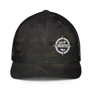 Puede incluir: Una gorra de camionero de camuflaje negro y verde con una brújula bordada en blanco y el texto "Let's Go North" dentro de la brújula. La brújula también presenta un contorno blanco de la península superior de Michigan.