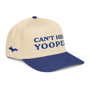Può includere: Un cappellino da baseball con corona color crema e visiera blu navy. Il testo "CAN'T HIDE YOOPER" è stampato in blu navy sul davanti. Una silhouette blu della Upper Peninsula del Michigan è sul lato.