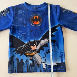 May include: Blue long-sleeved Batman shirt. The shirt features a graphic of Batman flying over a city skyline. The word "BATMAN" is printed vertically on the shirt. The shirt measures approximately 16 inches in length.