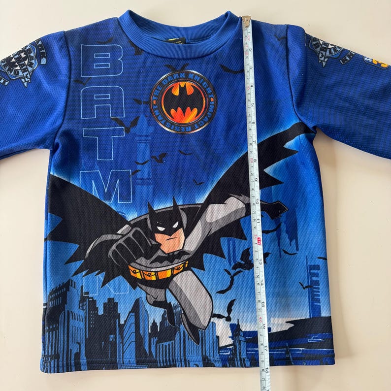 May include: Blue long-sleeved Batman shirt. The shirt features a graphic of Batman flying over a city skyline. The word "BATMAN" is printed vertically on the shirt. The shirt measures approximately 16 inches in length.