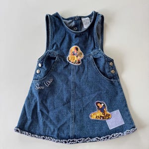 May include: A blue denim overall dress with two Snow White patches and the embroidered text "Snow White". The dress has two front pockets, button closures on the straps and sides, and a ruffled hem.
