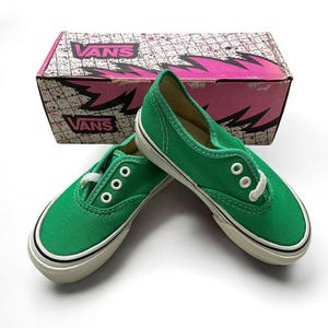 May include: A pair of green Vans trainers with white laces and white soles. The shoes are sitting on a white surface and are in front of a pink and white Vans shoebox.