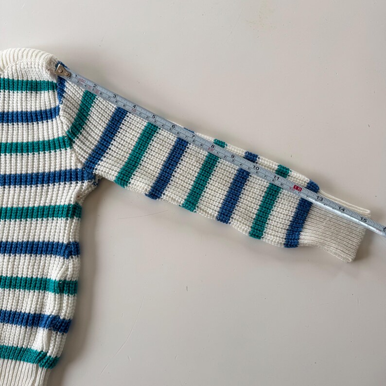 May include: A white, blue, and teal striped sweater sleeve is measured with a ruler. The sleeve is approximately 15 inches long. The sweater has a ribbed knit texture and a classic, nautical-inspired design.