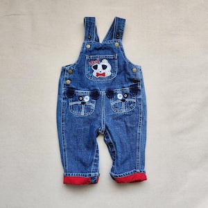 May include: Blue denim baby dungarees with a dog design. Features a dog face applique on the bib pocket and two dog face designs on the front pockets. The cuffs are red. Includes adjustable straps and button closures.