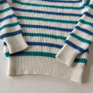 May include: Close-up of a striped sweater. The sweater features horizontal stripes in white, teal, and blue. The cuffs and bottom hem are solid white. The knitwear has a ribbed texture.