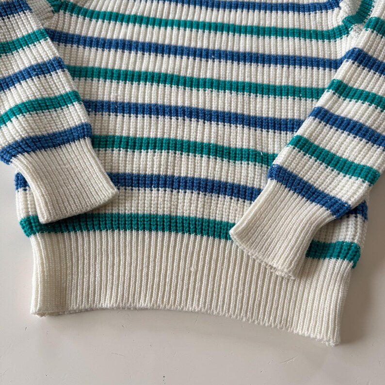 May include: Close-up of a striped sweater. The sweater features horizontal stripes in white, teal, and blue. The cuffs and bottom hem are solid white. The knitwear has a ribbed texture.