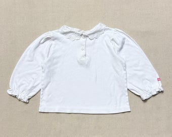 May include: White long-sleeved shirt featuring a lace collar and button fastening. The sleeves have a gathered cuff detail. A small pink tag is visible on the sleeve. The shirt is made of a soft, lightweight fabric.