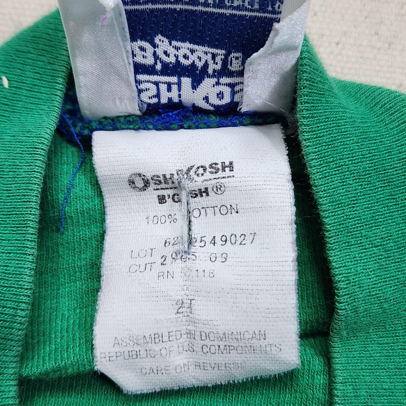May include: Close-up of a green toddler's shirt with a white tag. The tag reads "OSHKOSH B'GOSH" and "100% COTTON". Additional text includes "2T" and "ASSEMBLED IN DOMINICAN REPUBLIC OF U.S. COMPONENTS".