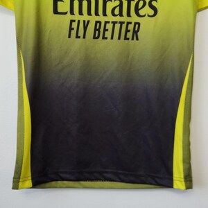 May include: A lime green and black gradient athletic jersey featuring the black text Emirates FLY BETTER printed on the chest. The shirt includes lime green mesh side panels and a smooth fabric finish throughout the main body.