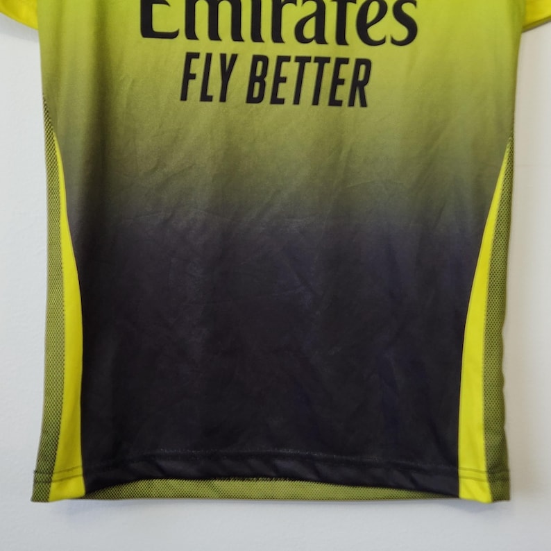 May include: A lime green and black gradient athletic jersey featuring the black text Emirates FLY BETTER printed on the chest. The shirt includes lime green mesh side panels and a smooth fabric finish throughout the main body.