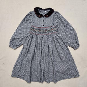 May include: A long-sleeved dress with a black and white gingham pattern. The dress features a black velvet collar, decorative embroidery, and a smocked bodice. The dress has a gathered skirt and is suitable for children.