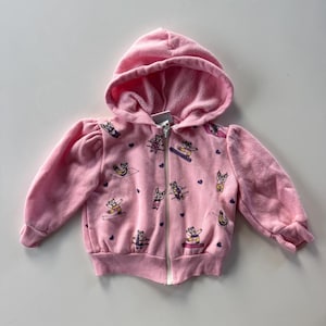 May include: A pink hooded sweatshirt with a full zipper closure. The sweatshirt features a pattern of cartoon bears engaged in various activities, such as yoga and skiing, along with small purple hearts. The sleeves have a puffy design.