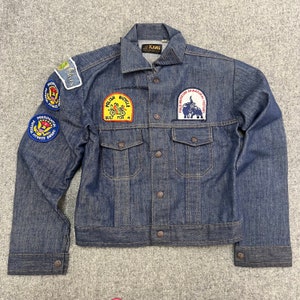Peut inclure: Une veste en jean bleu foncé avec plusieurs patchs cousus sur le devant. Les patchs comprennent un Polish Bicycle Built for 4, Field Museum of Natural History, Presidential Fitness Award et un patch avec le mot "Futures".