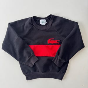 May include: Black sweatshirt with a red horizontal stripe across the chest. Features a red crocodile logo on the chest. The sweatshirt has a crew neck and long sleeves.