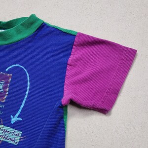 May include: A colorful short-sleeved shirt with a blue body, a purple sleeve, and a green collar. The shirt features a graphic with text and an arrow. The fabric has a ribbed texture.