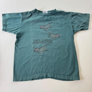 May include: A dark green short-sleeve t-shirt featuring a graphic print of three gray alligators with pink mouths. The text "NORTH CAROLINA AQUARIUMS" is printed in black lettering between the alligators on the front of the garment.