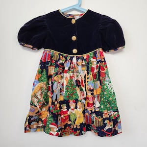 May include: A short-sleeved dress featuring a dark navy blue velvet bodice with three gold-toned buttons and a round neckline. The skirt is made of a dark navy blue fabric printed with a colorful pattern of teddy bears, Christmas trees, and gifts.