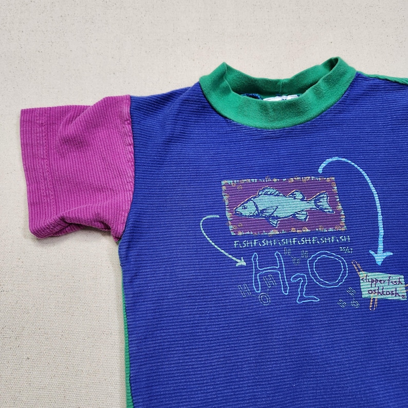 May include: A blue short-sleeved shirt with a green collar and a purple sleeve. The shirt features a fish graphic with the text "FISH FISH FISH FISH FISH FISH" and "H2O".