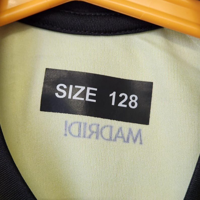 May include: A light yellow garment with a black collar and a black rectangular label featuring the white text SIZE 128. Below the label, the word MADRID is printed in a dark blue, inverted, and mirrored font on the fabric surface.
