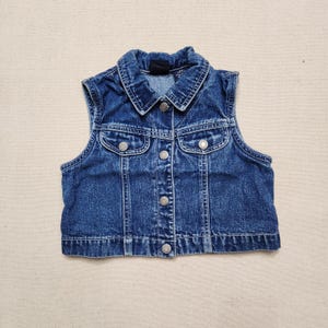 Könnte beinhalten: Eine dunkelblaue Jeansweste mit klassischem Kragen, einer durchgehenden Knopfleiste mit Metallknöpfen und zwei Brusttaschen mit geknöpften Klappen. Das ärmellose Kleidungsstück hat einen geraden Saum und durchgehend sichtbare Nähte.