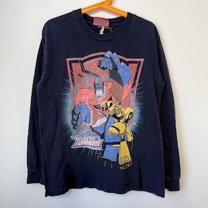 Könnte beinhalten: Ein dunkelblaues Langarm-T-Shirt mit einem Grafikdruck von Optimus Prime und Bumblebee aus Transformers. Das Design zeigt das Transformers-Logo, einen roten Autobot-Symbol-Hintergrund und blaue Gittermuster auf der Vorderseite des T-Shirts.