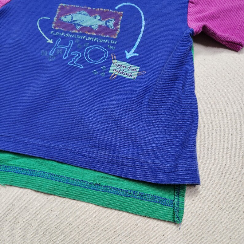 May include: A blue, green, and purple color-blocked t-shirt with a graphic design. The design includes a fish, the letters "H2O", and the words "dippo luk oshkosh". The shirt has a ribbed texture.