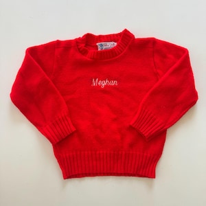 May include: A bright red sweater with long sleeves and ribbed cuffs and hem. The word "Meghan" is embroidered in white script across the chest. The sweater has a crew neck and a small label at the neckline.