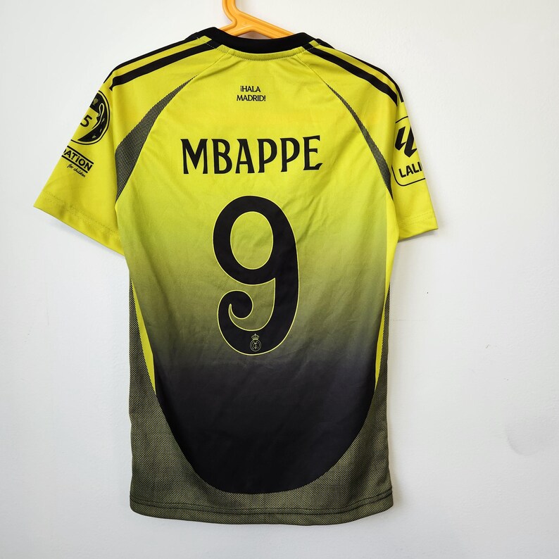 May include: A yellow and black soccer jersey featuring the name MBAPPE and the number 9 printed in black on the back. The shirt includes the text &iexcl;HALA MADRID! near the collar, along with circular patches on the sleeves and black side panels.