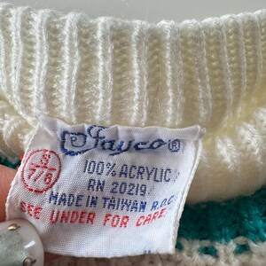 May include: Close-up of a white and teal knit sweater with a tag. The tag reads "Jayco" and "100% Acrylic." The tag also indicates the sweater was made in Taiwan. The sweater has a ribbed knit collar.