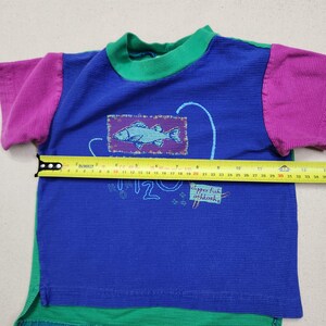 May include: A blue, green, and purple short-sleeved shirt with a fish graphic. The shirt has a green collar and hem, purple sleeves, and a fish illustration on the front. Text on the shirt reads "How 12" and "shipper fish oshkosh".