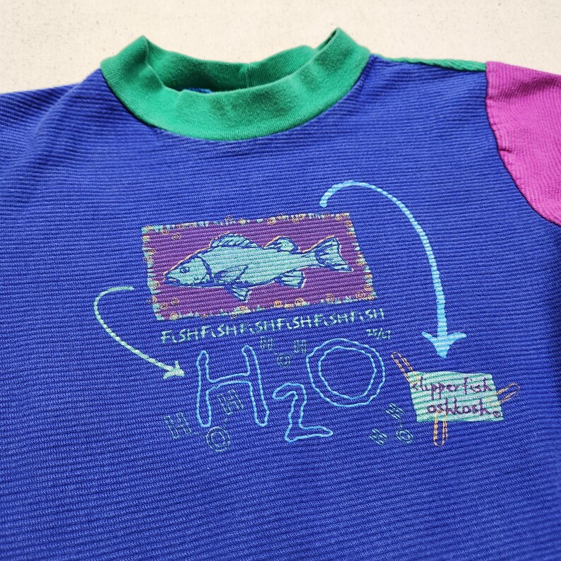 May include: Blue long-sleeved shirt with a green collar and purple sleeve. The shirt features a fish graphic inside a purple rectangle, with the words "FISH FISH FISH FISH FISH" below it. The shirt also has the word "H2O" and the words "slipper fish oshkosh" printed on it.
