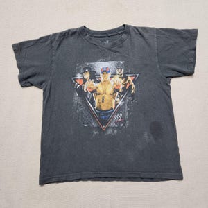 May include: A charcoal gray short-sleeve t-shirt featuring a graphic print of three professional wrestlers inside a triangular frame. The center figure wears a blue cap, while the WWE logo is printed in the bottom right corner of the graphic.