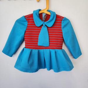 May include: A long-sleeved top featuring a red bodice with horizontal blue stripes, a solid blue collar, and a matching blue tie-shaped accent. The garment includes a pleated blue peplum hem and is displayed on a yellow plastic hanger.