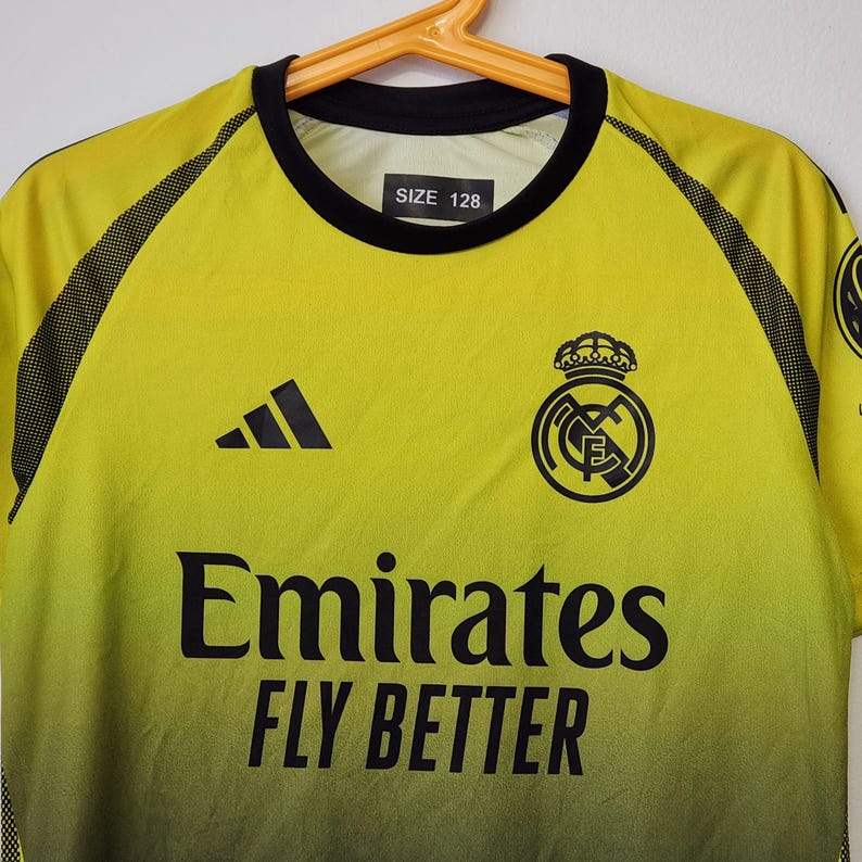 May include: A yellow and black gradient soccer jersey featuring the Real Madrid crest, a black Adidas logo, and the text Emirates FLY BETTER in black. The shirt has a black crew neckline and a size 128 label printed on the inside of the collar.
