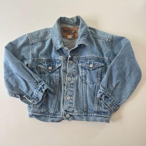 May include: A light blue denim jacket featuring a classic collar, long sleeves with buttoned cuffs, and a front button closure. The garment includes two chest pockets with buttoned flaps and a brown leather-like brand patch on the inner neck.
