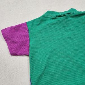 May include: A short-sleeved shirt with a green body and a purple sleeve. The shirt has a ribbed texture and a high neckline. The sleeve is a solid purple color.