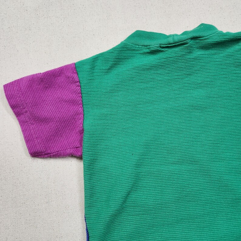 May include: A short-sleeved shirt with a green body and a purple sleeve. The shirt has a ribbed texture and a high neckline. The sleeve is a solid purple color.