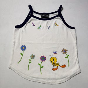 May include: A white tank top with dark blue trim featuring a graphic of Tweety Bird, colorful flowers, and small dragonflies. The inner neck label displays the text Looney Tunes Collection. The design includes a yellow bird, green stems, and petals in blue, purple, orange, and yellow.