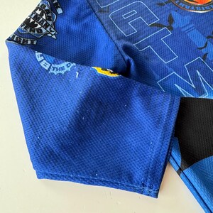 May include: Close-up of a blue athletic jersey with a mesh texture. The jersey features white and yellow graphics, including text and a circular emblem. The sleeve is visible, with a black panel on the side. The text "THE" is also visible.