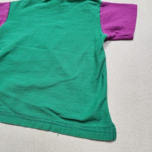 May include: A green and purple short-sleeved shirt. The body of the shirt is green with horizontal ribbed texture. The sleeves are purple. The shirt is made of a soft, ribbed fabric.