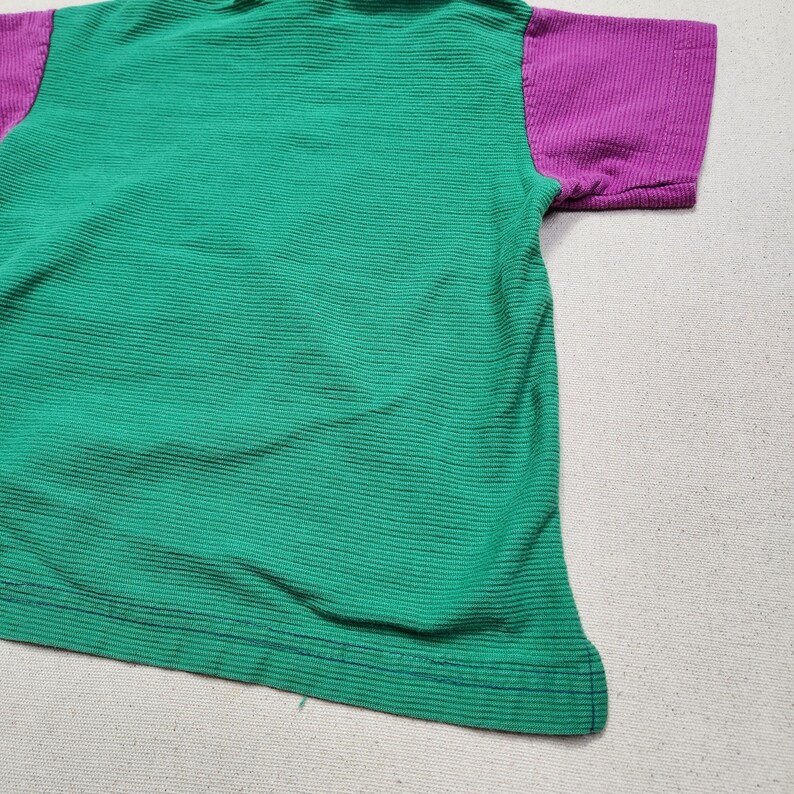 May include: A green and purple short-sleeved shirt. The body of the shirt is green with horizontal ribbed texture. The sleeves are purple. The shirt is made of a soft, ribbed fabric.