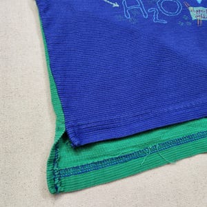 May include: Close-up of a blue and green t-shirt. The blue section has the text "H2O" and a small graphic. The shirt has a ribbed texture. The bottom hem is green with blue stitching.