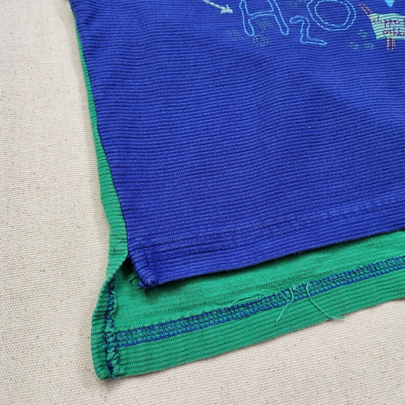 May include: Close-up of a blue and green t-shirt. The blue section has the text "H2O" and a small graphic. The shirt has a ribbed texture. The bottom hem is green with blue stitching.