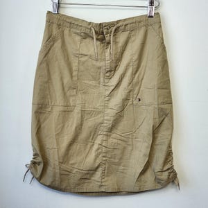 Puede incluir: Una falda color caqui hasta la rodilla con cintura elástica y cordón, dos bolsillos delanteros y cordones ajustables en el dobladillo inferior. Un pequeño parche con el logotipo de Tommy Hilfiger es visible en el bolsillo delantero izquierdo.