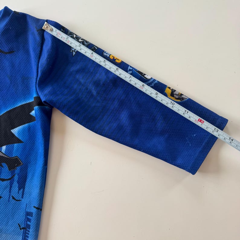 May include: A blue long-sleeved garment with a black graphic design. A measuring tape shows the sleeve is approximately 14 inches long. The garment has a textured appearance.