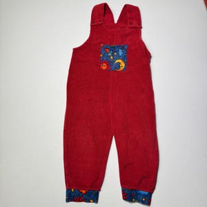 May include: Red children's overalls with a pocket featuring a colorful space-themed design of rockets, suns, and moons. The cuffs are blue with a similar space pattern. The overalls have adjustable straps with button closures.