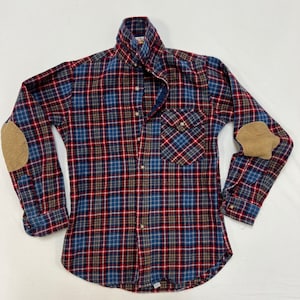 Peut inclure: Une chemise en flanelle à carreaux bleus, rouges et bruns avec des empiècements aux coudes. La chemise a une fermeture boutonnée à l'avant et un col pointu.
