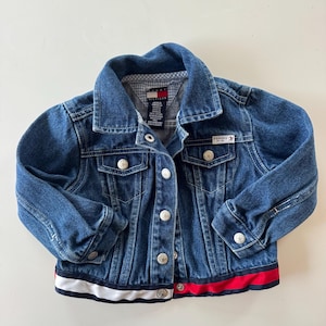 以下が含まれることがあります： シルバーのボタンと赤、白、青のストライプの裾が付いた青いデニムジャケット。 ジャケットには2つの胸ポケットと小さなロゴパッチが付いています。 ジャケットはクラシックスタイルです。