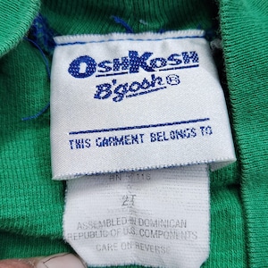 May include: Close-up of a green garment tag with the OshKosh B'gosh logo in blue. The tag reads "THIS GARMENT BELONGS TO" and includes size "2T". The garment is assembled in the Dominican Republic with U.S. components.