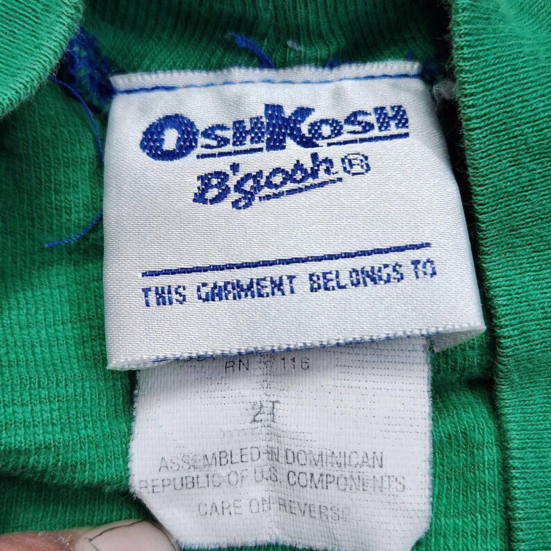 May include: Close-up of a green garment tag with the OshKosh B'gosh logo in blue. The tag reads "THIS GARMENT BELONGS TO" and includes size "2T". The garment is assembled in the Dominican Republic with U.S. components.